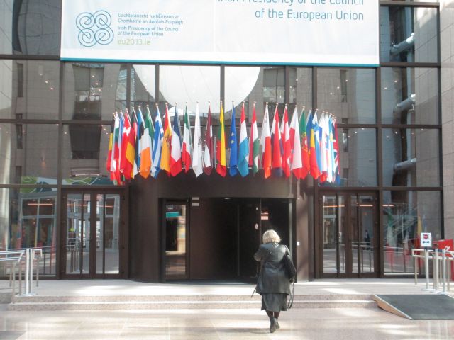 Foto av ingången till EUs rådsbyggnad. En person är på väg in. Många flaggor hänger över ingången. 